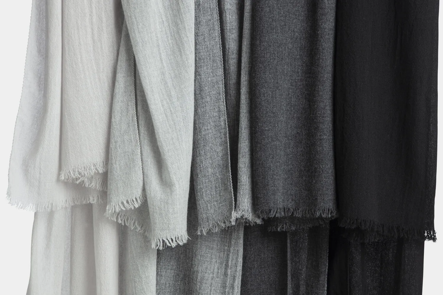 Collection of pure cashmere scarves in soft neutral tones, ranging from light grey to charcoal and black, draped to showcase their lightweight texture and delicate fringed edges.