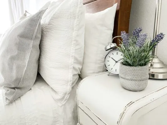 Layered white bedding with striped and textured pillows beside a bedside table with flowers and an alarm clock.
